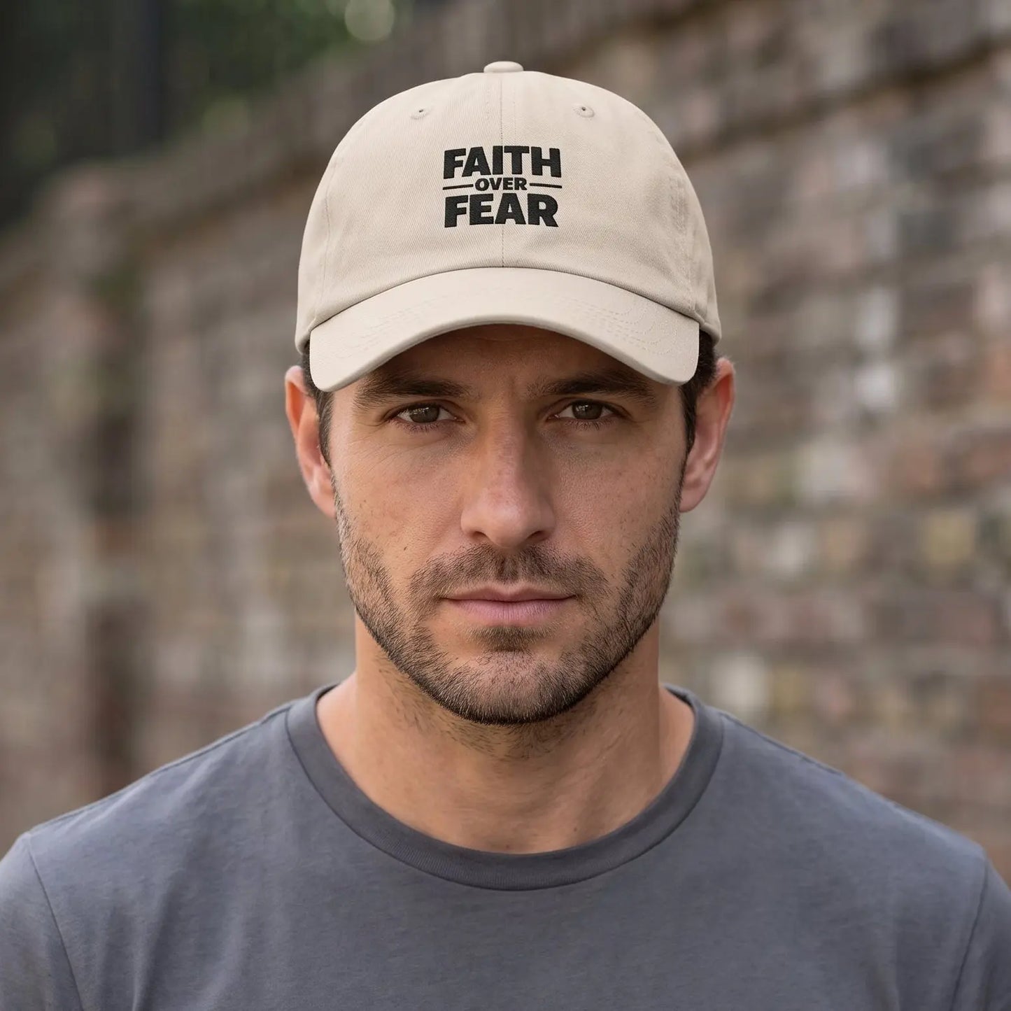 A man wearing a beige baseball cap with the black text 'FAITH OVER FEAR' and a dark gray t-shirt, standing in front of a blurred brick wall.