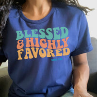 May include: A navy blue t-shirt with the text &#39;Blessed & Highly Favored&#39; in rainbow colors. The text is in a retro font style. The text &#39;Luke 1:28&#39; is printed in a smaller font below the main text.