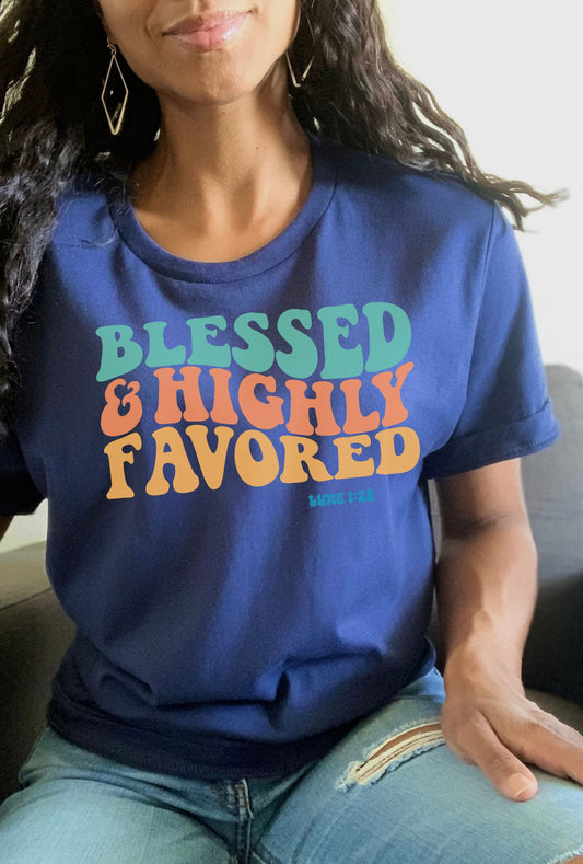 May include: A navy blue t-shirt with the text &#39;Blessed & Highly Favored&#39; in rainbow colors. The text is in a retro font style. The text &#39;Luke 1:28&#39; is printed in a smaller font below the main text.