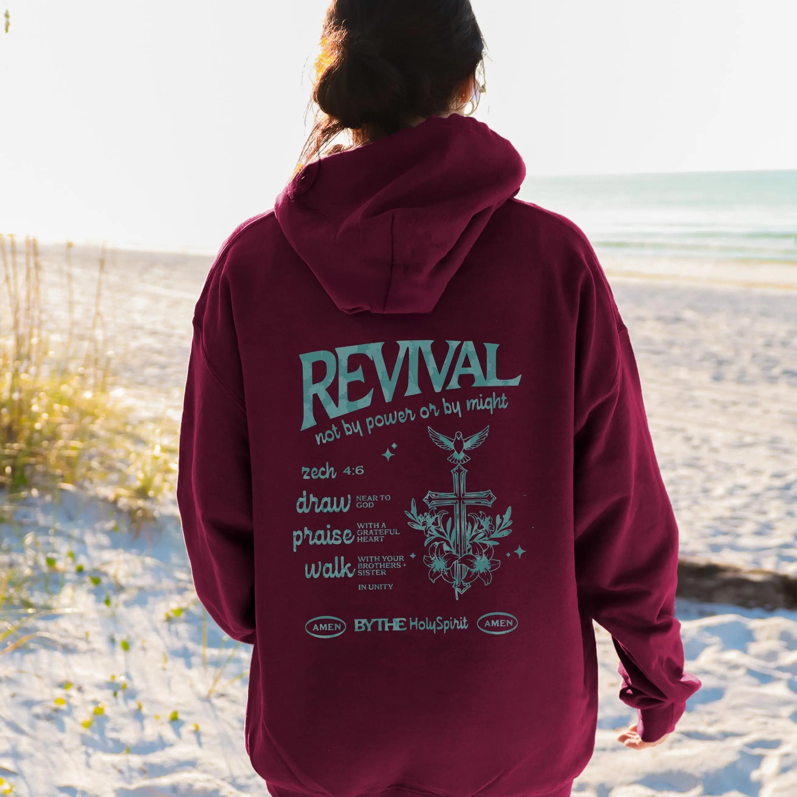 May include: A maroon hoodie with the word "REVIVAL" in teal script on the back, along with the text "not by power or by might" and other religious text. The hoodie is being worn on a beach.